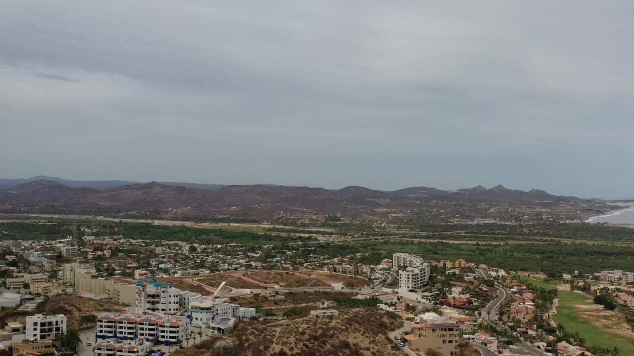 Second aerial frame in Los Cabos showing hillside neighborhoods, pale roads, green fairway-like areas, layered mountains, a distant strip of water, and no visible people.