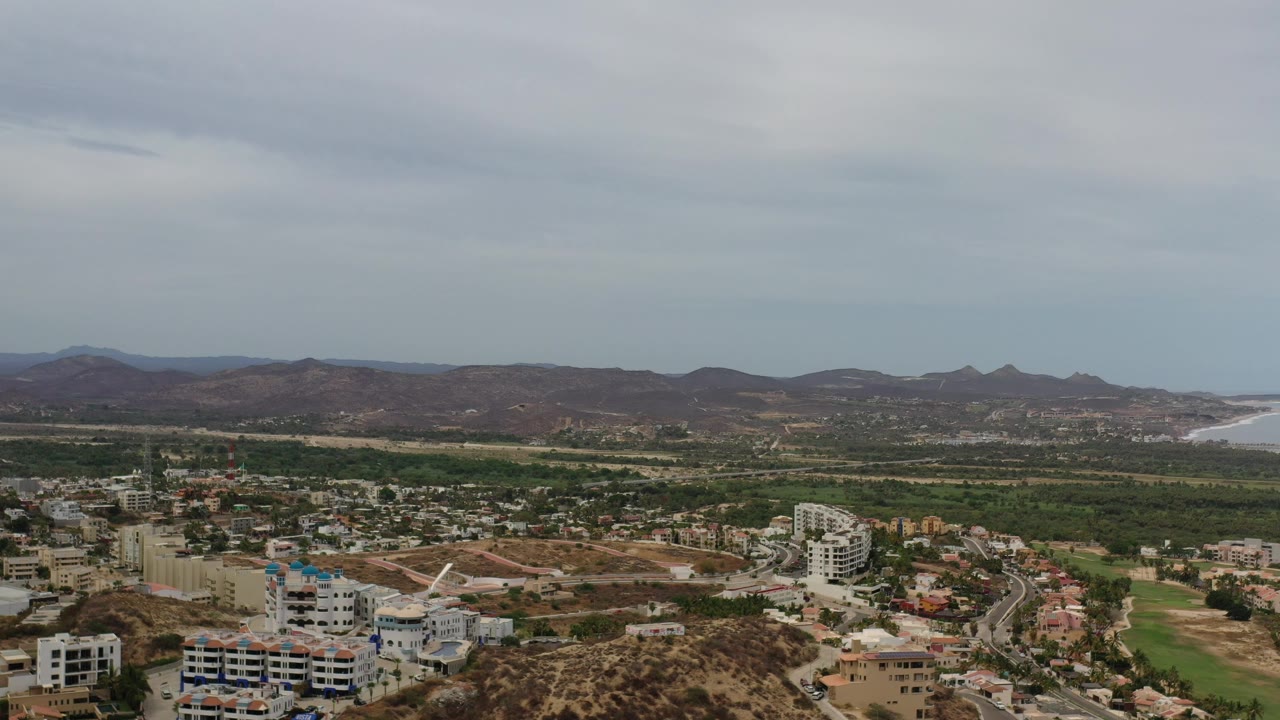 Aerial Los Cabos frame with compact rooftops and white buildings below, a broad green band through the midground, mountain ridges behind it, distant coastline, and no visible people.