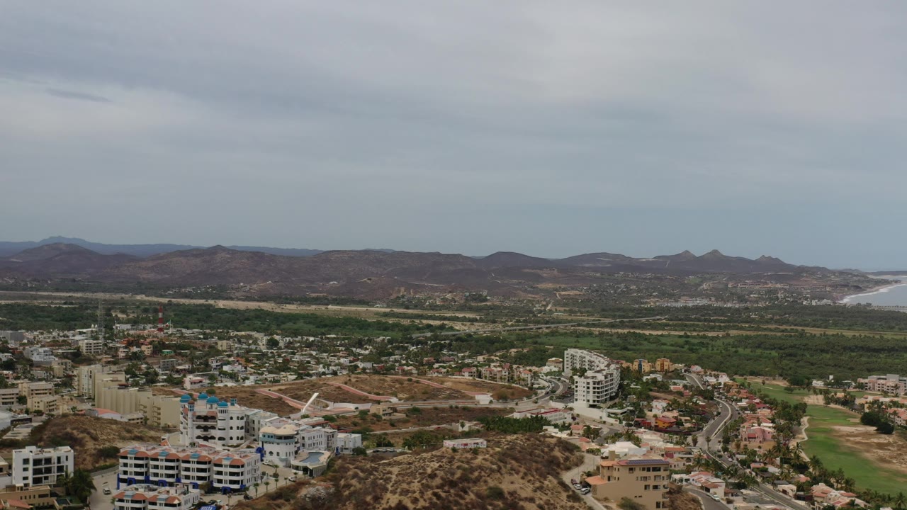 Aerial view of urban blocks, green ground, arid hills, mountains, and distant water.