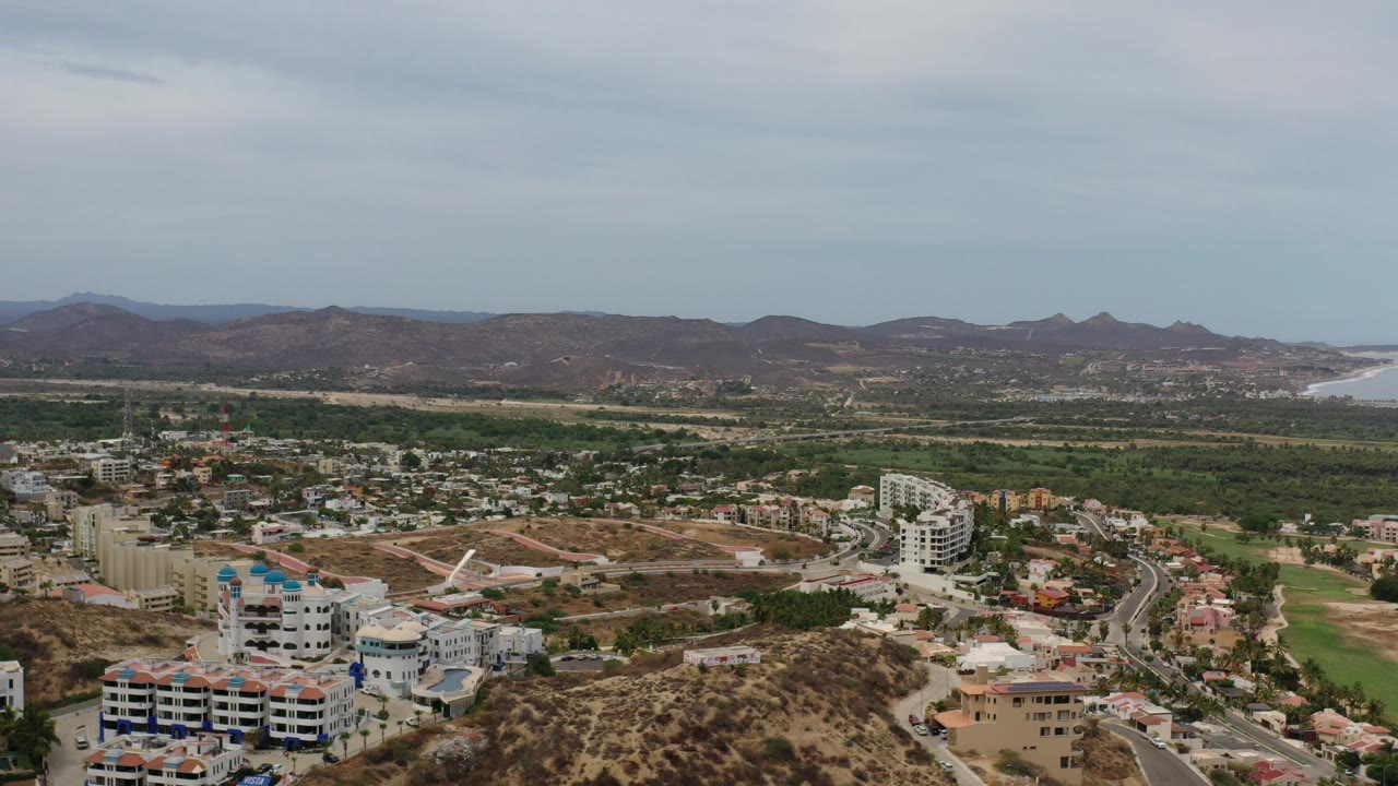 Opening aerial frame over Los Cabos, Mexico with white buildings in the foreground, curving roads, green grounds, distant mountains, an ocean inlet, and no visible people.