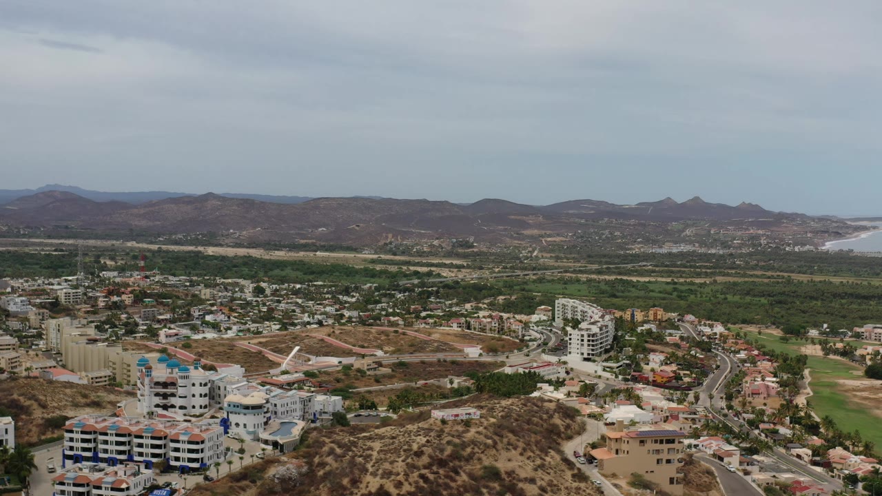 Aerial Los Cabos frame with a white multi-story building near the lower left, roads winding through developed blocks, low brown mountains, ocean beyond, and no visible people.