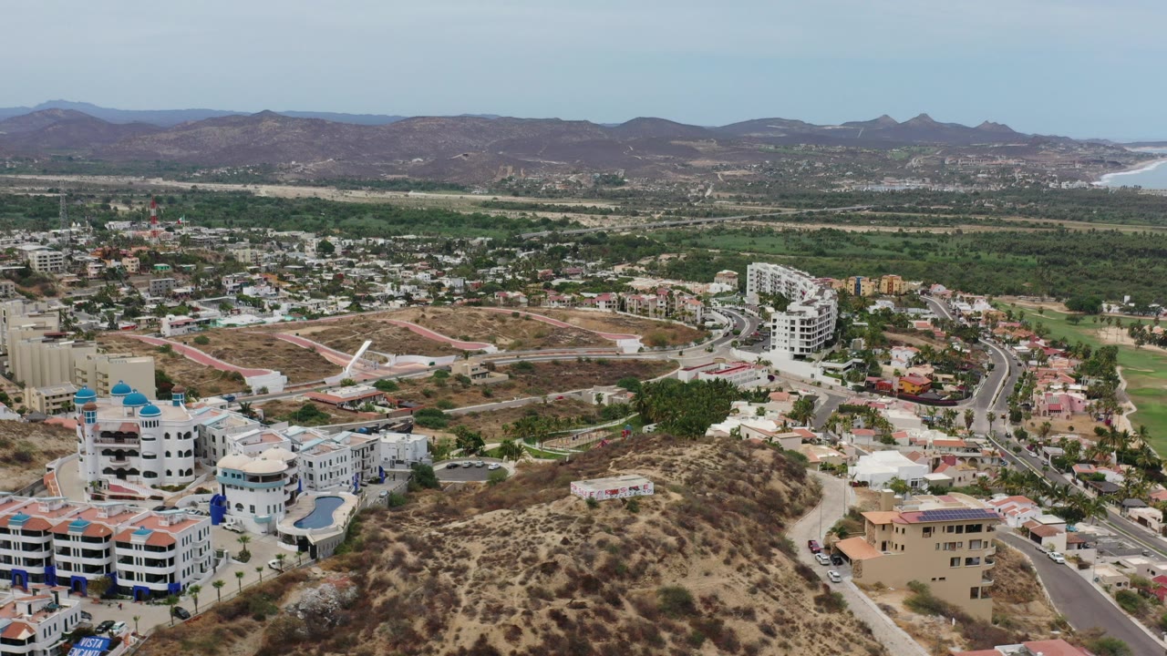 Wide aerial view in Los Cabos, Mexico, looking over a dry scrub hillside toward white and tan buildings, curving streets, green lowlands, distant mountains, and no visible people.
