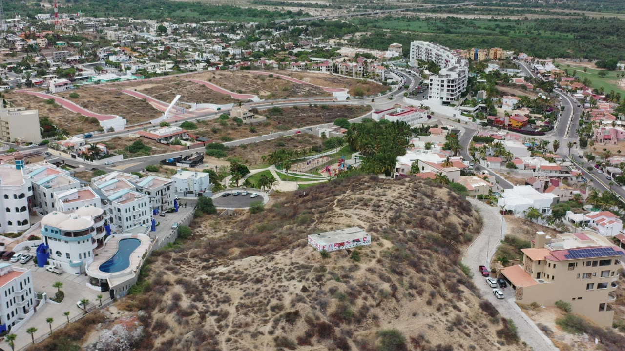 Opening aerial frame over Los Cabos, Mexico, with a scrubby dry hill in the foreground, resort-style buildings, curved roads, parked cars, and a blue pool, with no visible people.