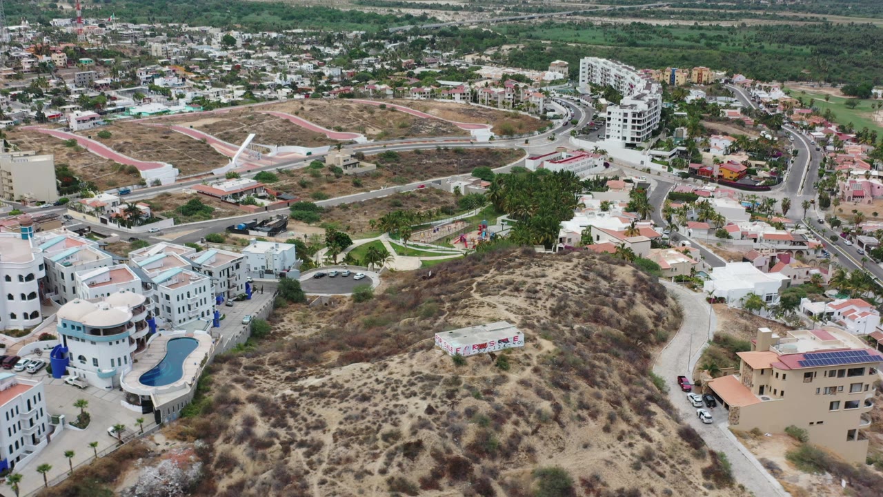 Aerial frame over Los Cabos, Mexico, showing the dry brush-covered slope centered lower in frame, a white rounded building, a blue swimming pool, nearby roads, and no visible people.