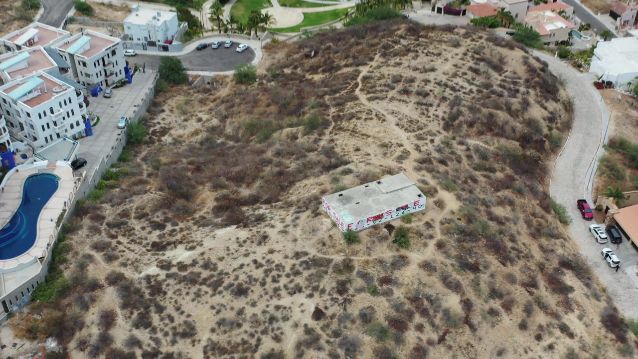 Aerial view of a flat-roofed house on a dry hillside near villas and a pool.