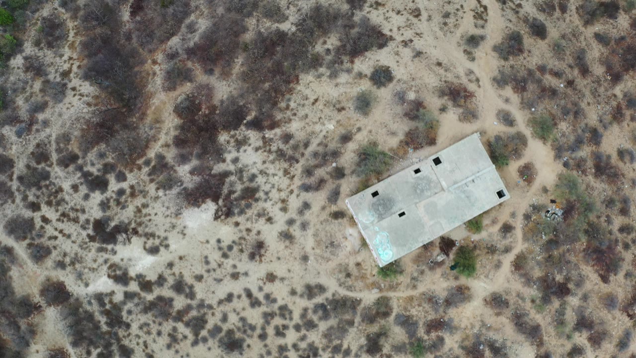 Overhead Los Cabos view with sandy scrubland, faint dirt paths, and a pale rectangular concrete building near the center, with no visible people.