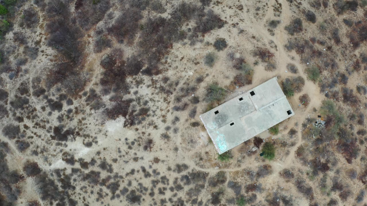 Aerial Los Cabos frame showing the concrete building slightly right of center amid beige dirt, dark brush patches, and narrow footpaths, with no visible people.