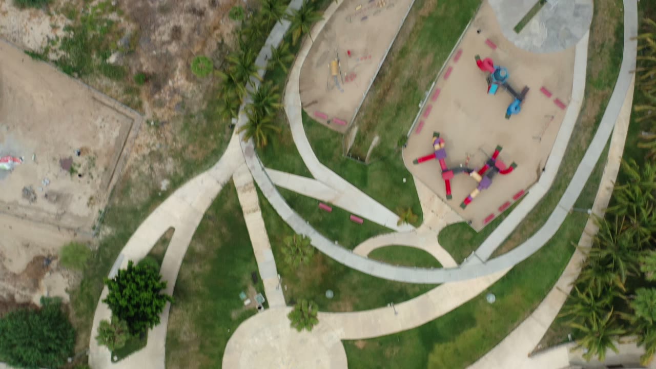 Overhead frame in Los Cabos, Mexico with a circular path layout, trimmed grass, palms, sandy lots, and red and blue playground equipment with no visible people.