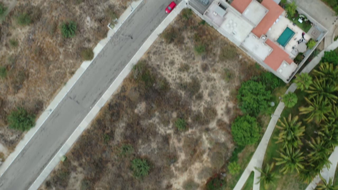 Overhead Los Cabos residential frame with a paved road, a white house with terracotta roof sections, a turquoise pool, palm trees, sidewalks, and dry lots, with no visible people.