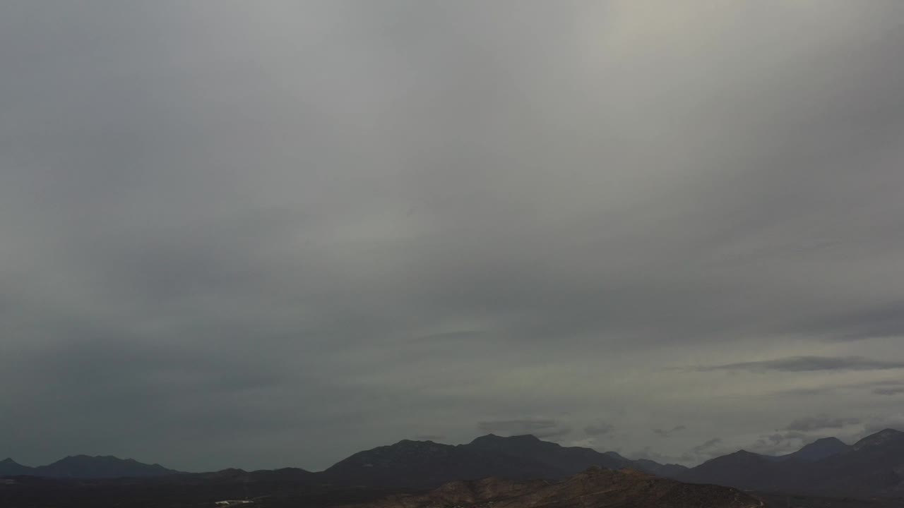 Los Cabos aerial frame with dark mountain ridges across the lower third, a large overcast sky above, and no visible people.
