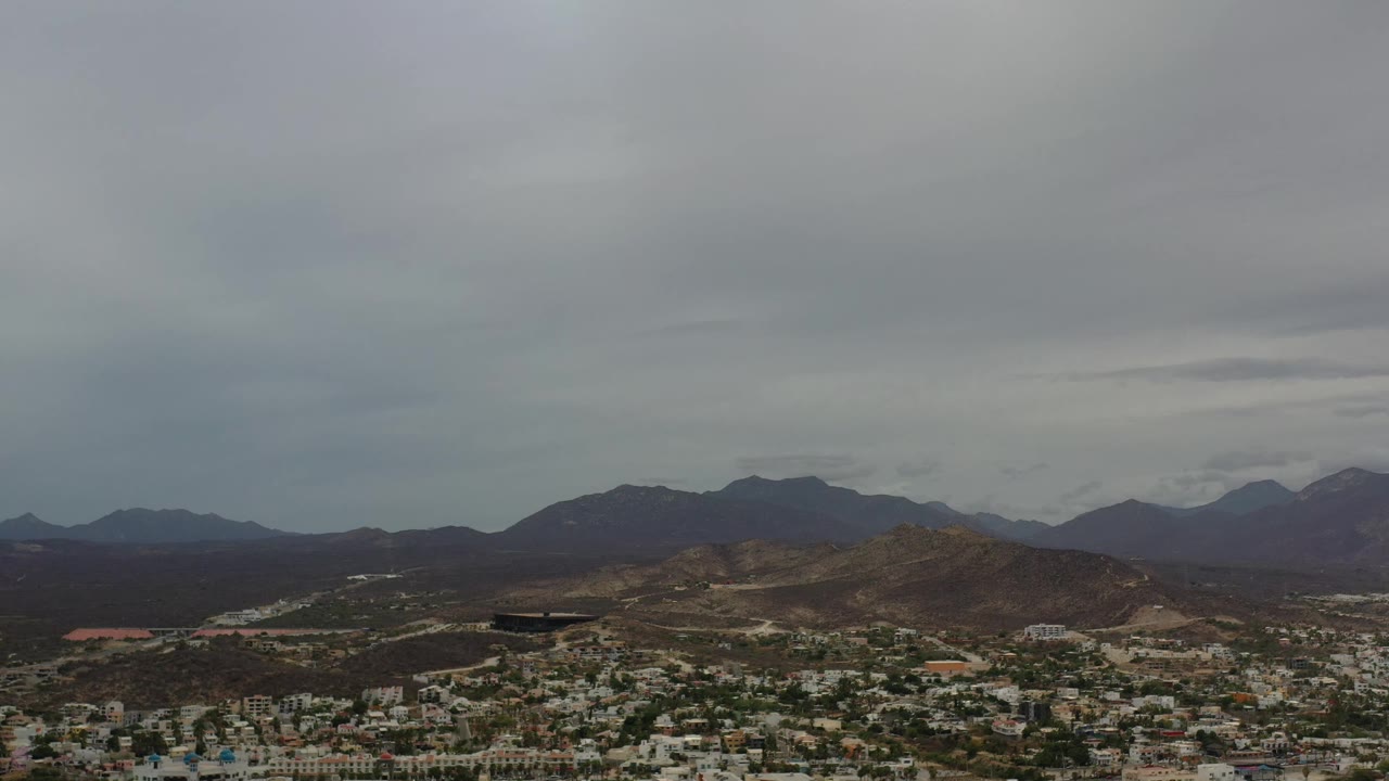 Early frame from the Los Cabos aerial view, with clustered buildings along the lower edge, dark mountain slopes in the distance, and no visible people.