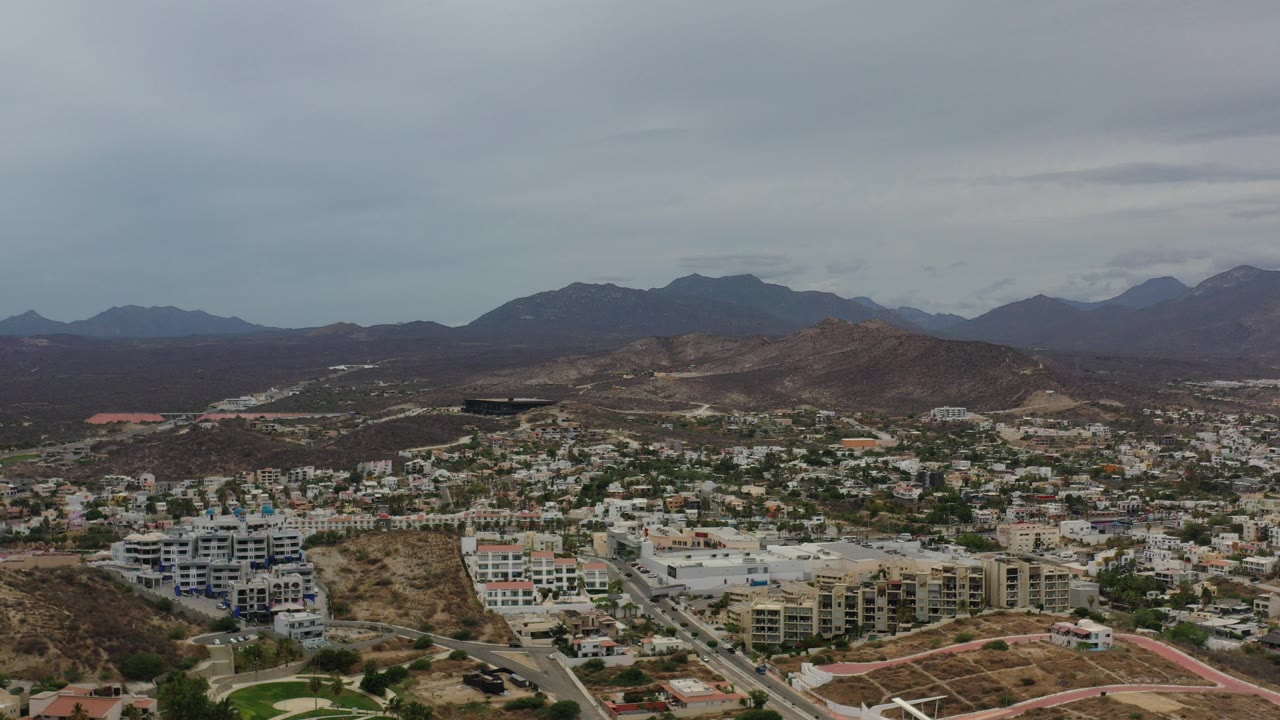 Aerial view of dry hills, city buildings, roads, and distant mountains under gray sky.