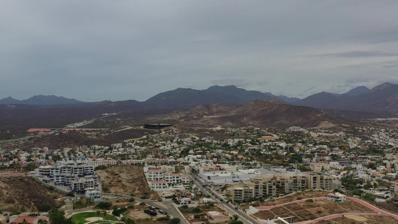 Aerial Los Cabos view with clustered white and beige buildings below, brown undeveloped slopes in the center, hazy mountains in the distance, and no visible people.