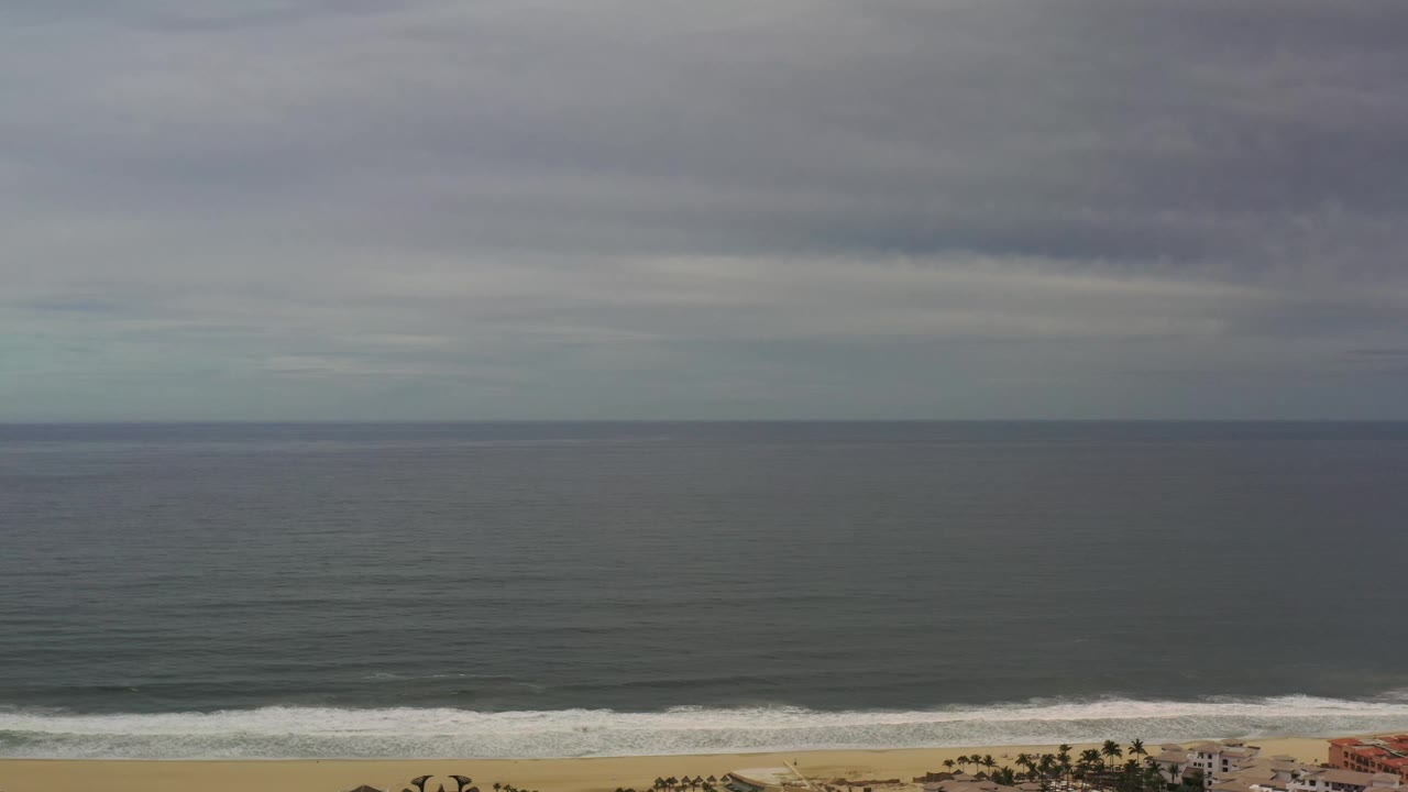 Los Cabos ocean view at 0:01 with the horizon centered, darker cloud bands across the sky, a thin line of breaking surf, beach sand, distant palms and buildings, and no visible people.