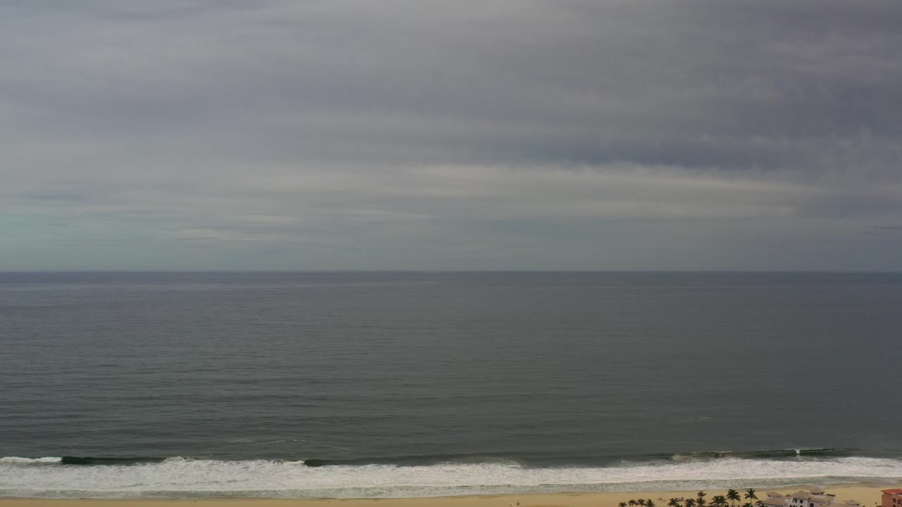 Los Cabos ocean horizon under a dense gray cloud layer, with dark water, a narrow sandy shoreline, small white surf, and no visible people.