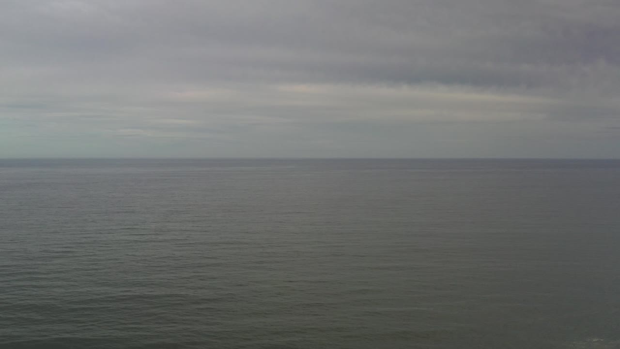 Wide aerial view over the gray-green ocean in Los Cabos, Mexico, with a flat horizon, layered cloud cover, and no visible people.