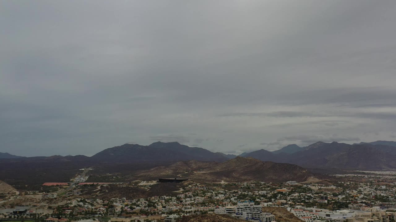 Los Cabos appears from an elevated view, with pale buildings at the bottom of frame, brown hills across the middle, distant mountains, and no visible people.