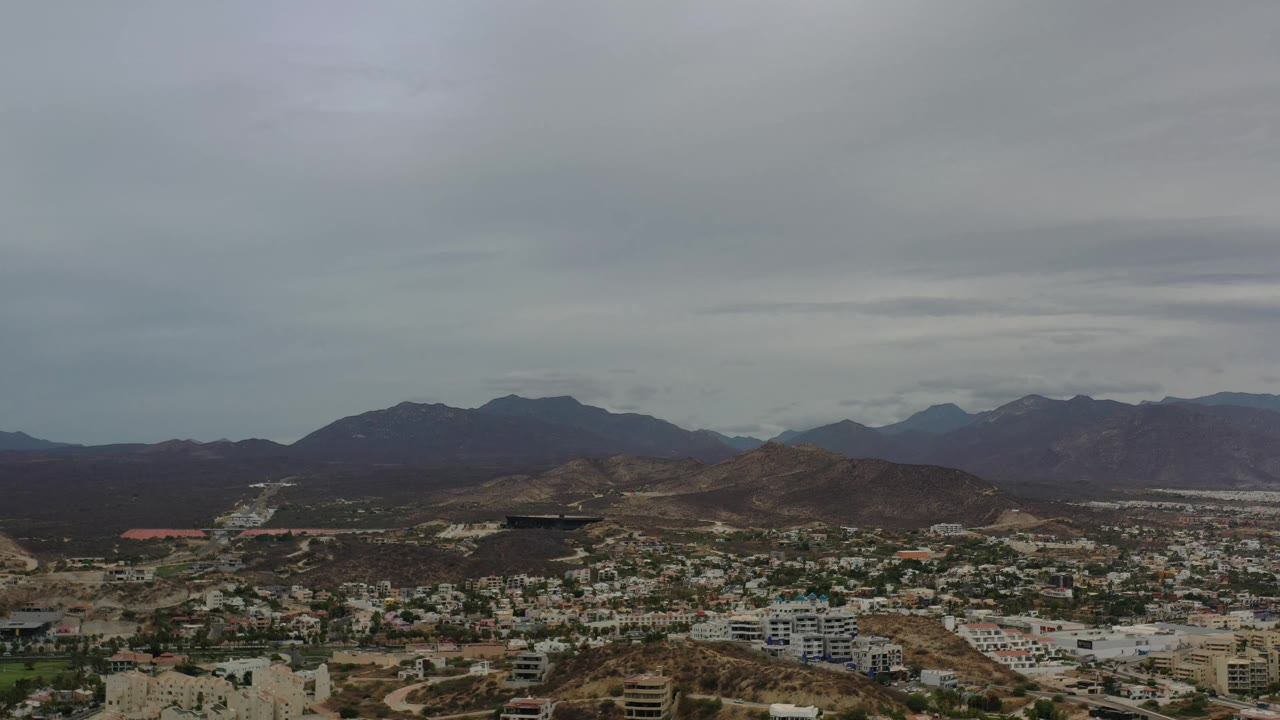 Aerial view in Los Cabos, Mexico, showing clustered buildings in the foreground, brown hills across the middle distance, dark mountains beyond, and no visible people.