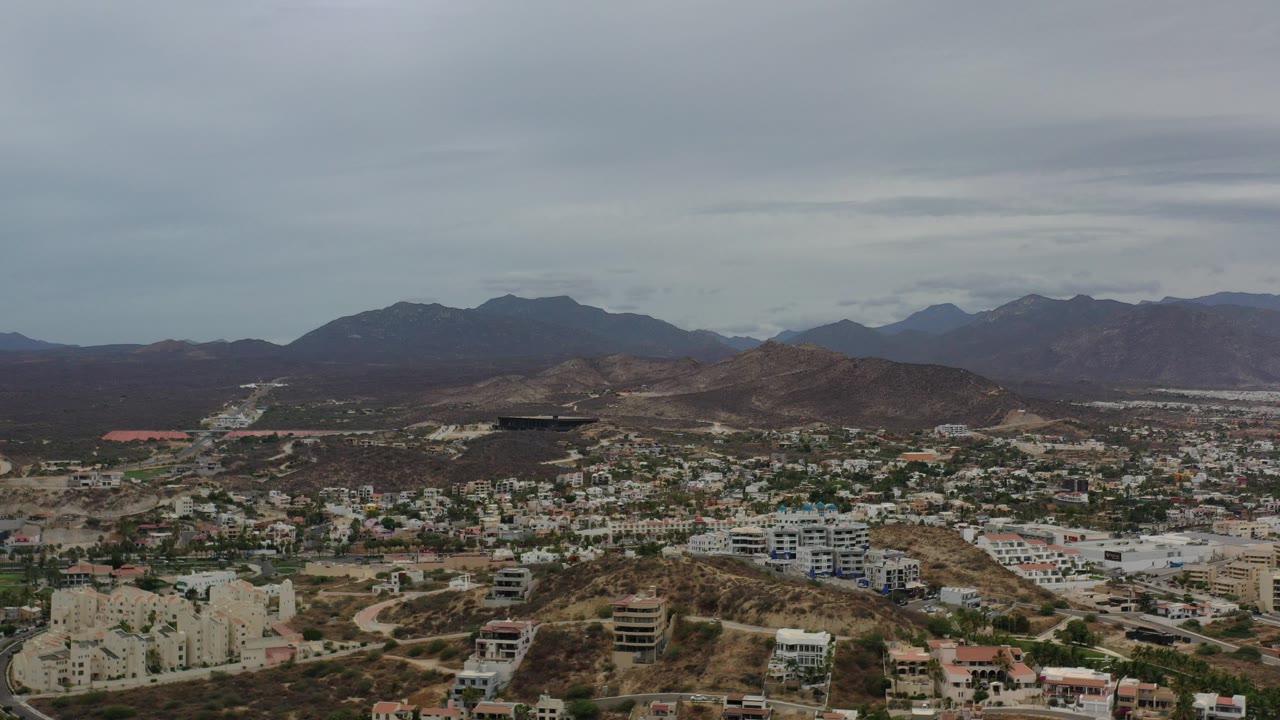 Opening aerial frame over Los Cabos, Mexico, showing hillside neighborhoods, winding roads, dry terrain, distant mountains, and no visible people.