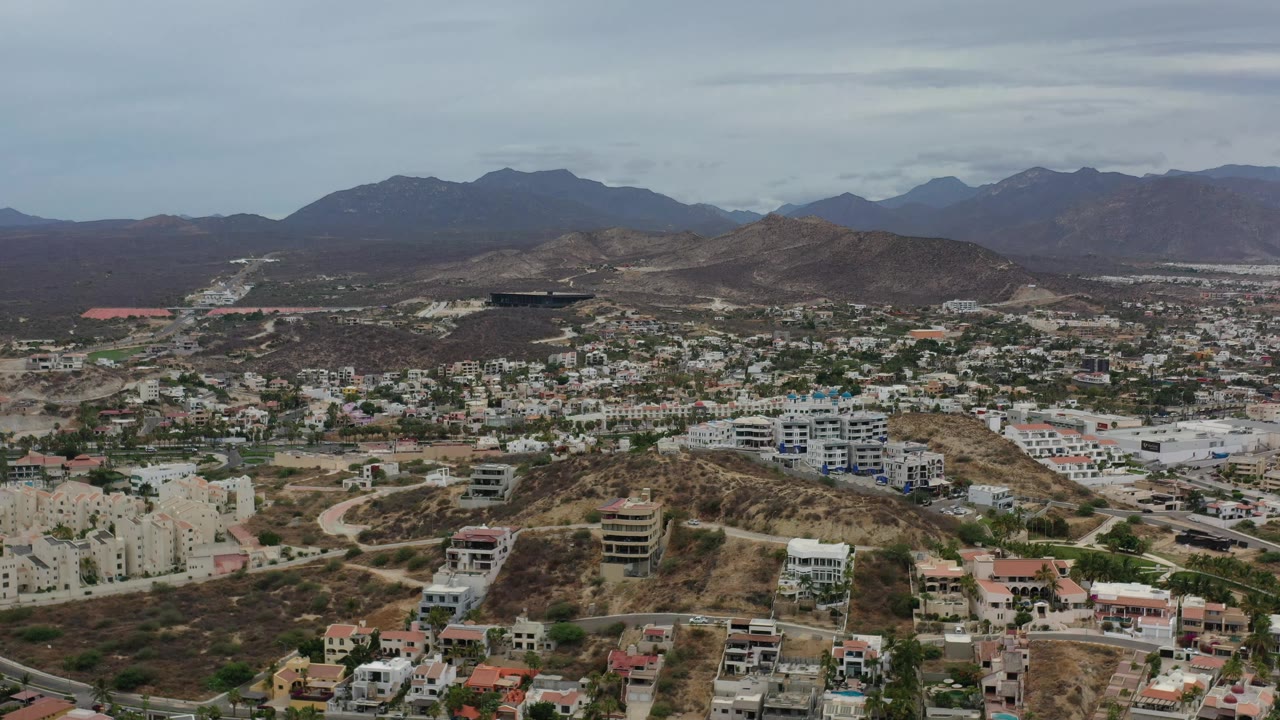 Early frame over Los Cabos, Mexico with white apartment blocks and homes spread across tan hills, a road grid in the foreground, dark mountain ridges beyond, and no visible people.