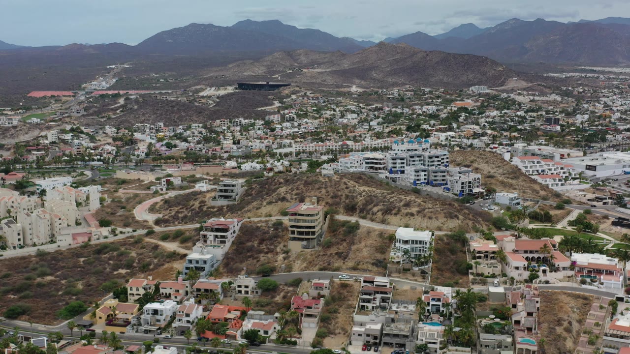Los Cabos aerial view one second in, showing clustered white buildings, brown hillside lots, curved roads, dark mountain ridges, and no visible people.