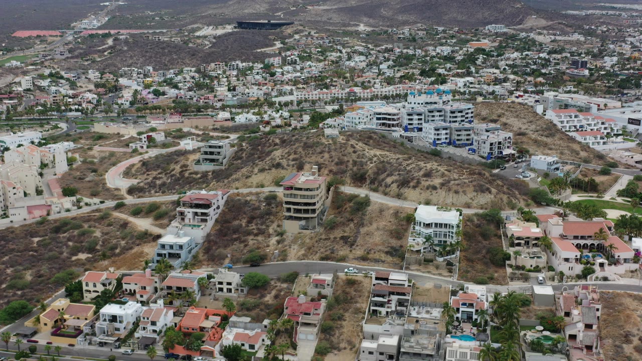 Aerial view over Los Cabos hillside homes, winding roads, dry scrub terrain, and distant residential blocks with no visible people.