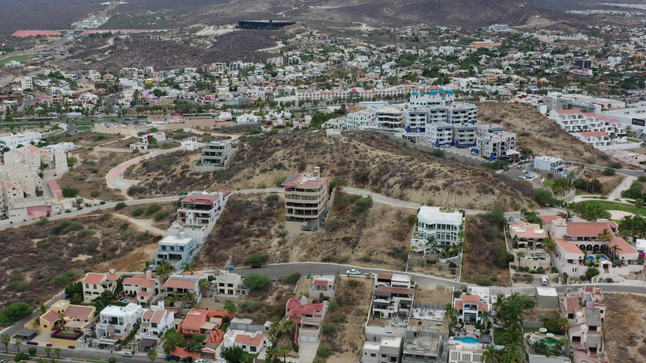 Aerial frame in Los Cabos showing a rounded dry hill, large white apartment-like buildings, villas, roads, and no visible people.