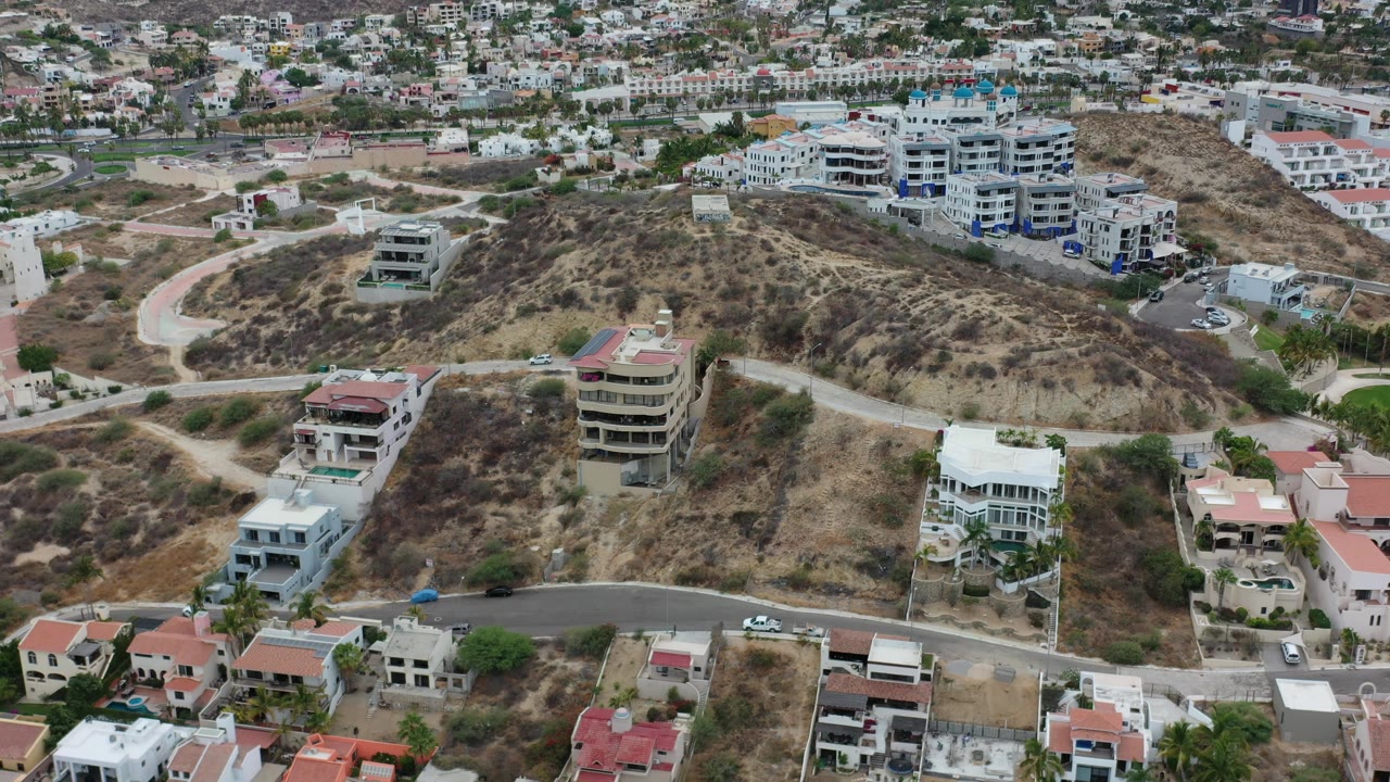 The Los Cabos aerial frame centers dry hillside lots, multi-story homes, winding roads, and a cluster of pale apartment buildings, with no visible people.