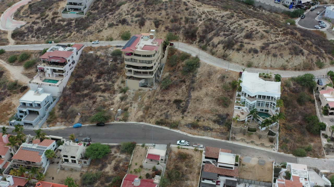 Overhead frame of modern homes on a dry hillside in Los Cabos, Mexico, with curving paved roads, parked vehicles, scrub vegetation, and no visible people.