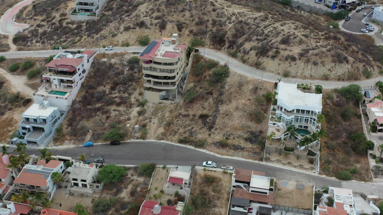 Overhead Los Cabos residential view with a round-front multi-story house near center, neighboring white homes, arid brush, road switchbacks, and no visible people.