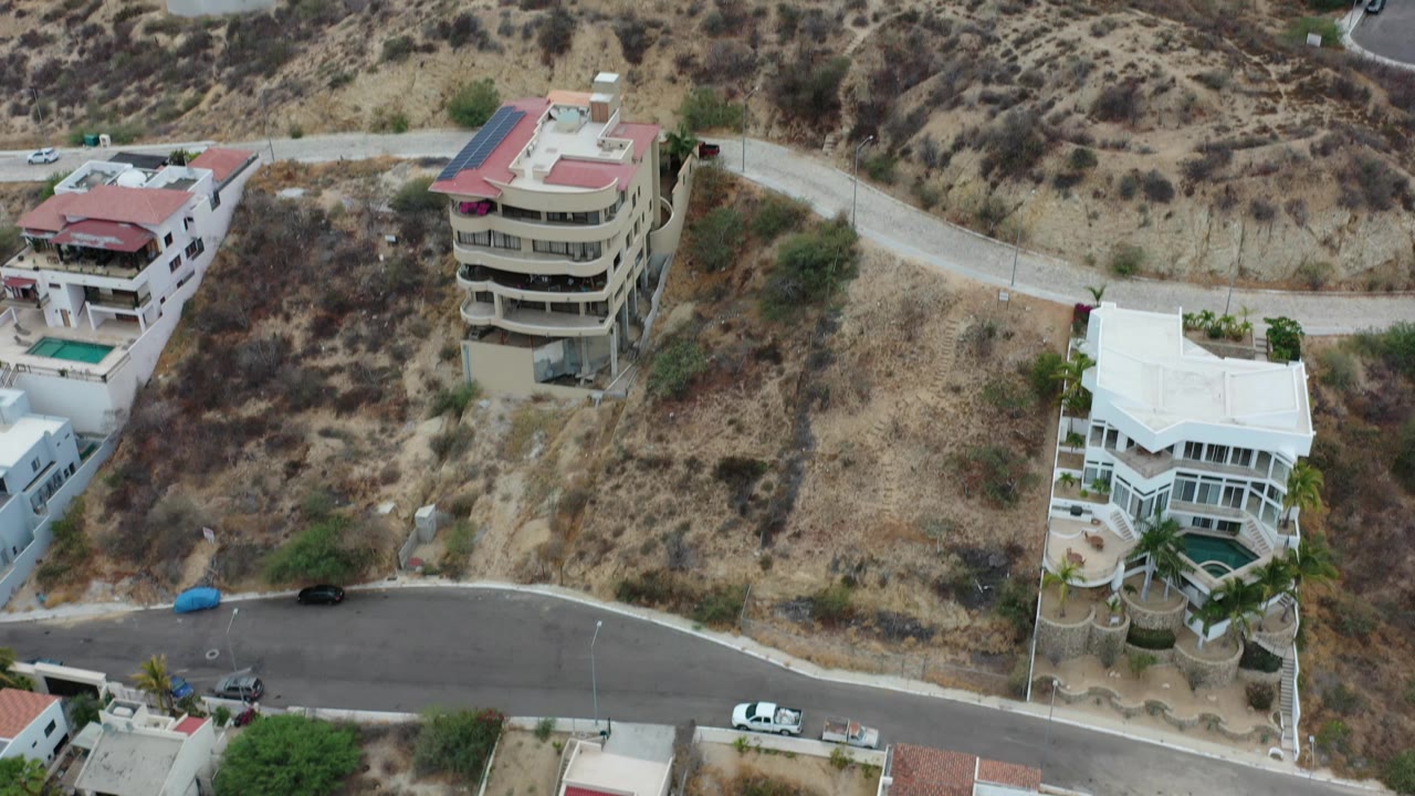 Drone view over Los Cabos, Mexico showing hillside homes, a curving paved road, dry scrub slopes, parked cars, and no visible people at the opening moment.