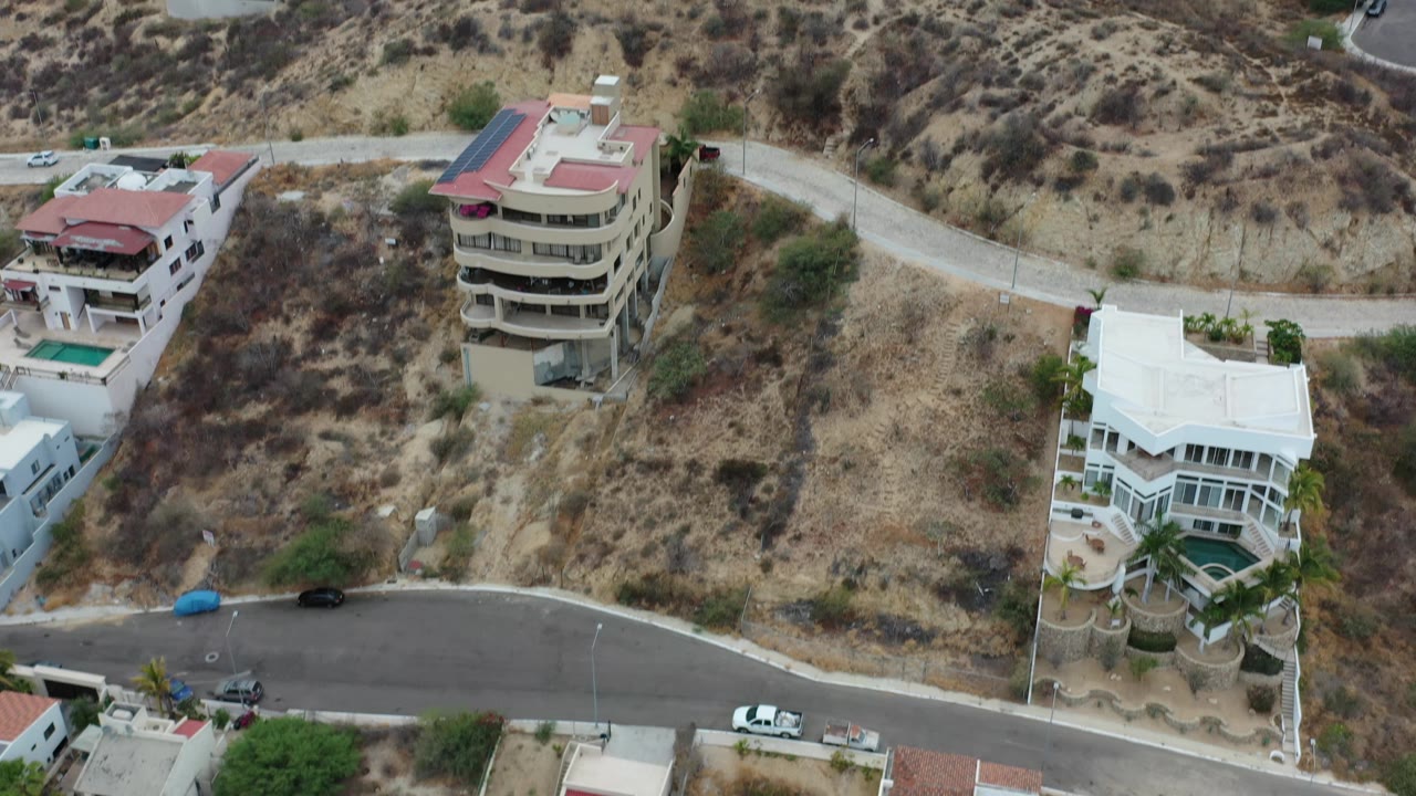 Drone frame over Los Cabos, Mexico with a beige multi-story building, white hillside house, narrow road, scrubby slope, parked cars, and no visible people.