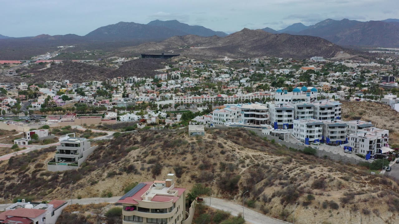 Opening aerial view in Los Cabos, Mexico, showing white hillside apartment blocks, scattered homes, dry terrain, distant mountains, and no visible people.
