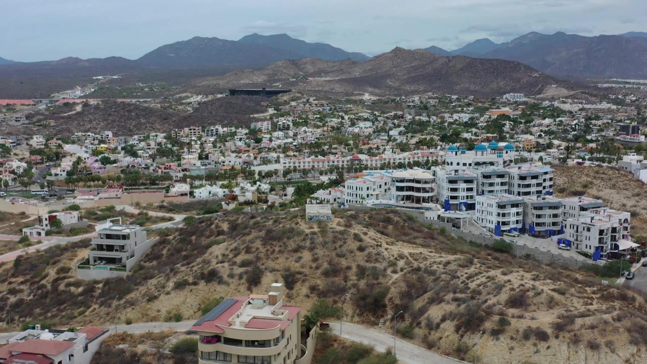 Los Cabos aerial frame with a large white multi-story building at right, low homes across the valley, dry hills in front, and no visible people.