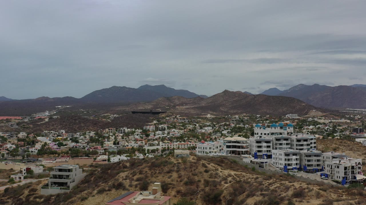 Wide elevated view in Los Cabos, Mexico of scrubby hills, white residential buildings, scattered houses, distant dark mountains, and a gray sky, with no visible people.