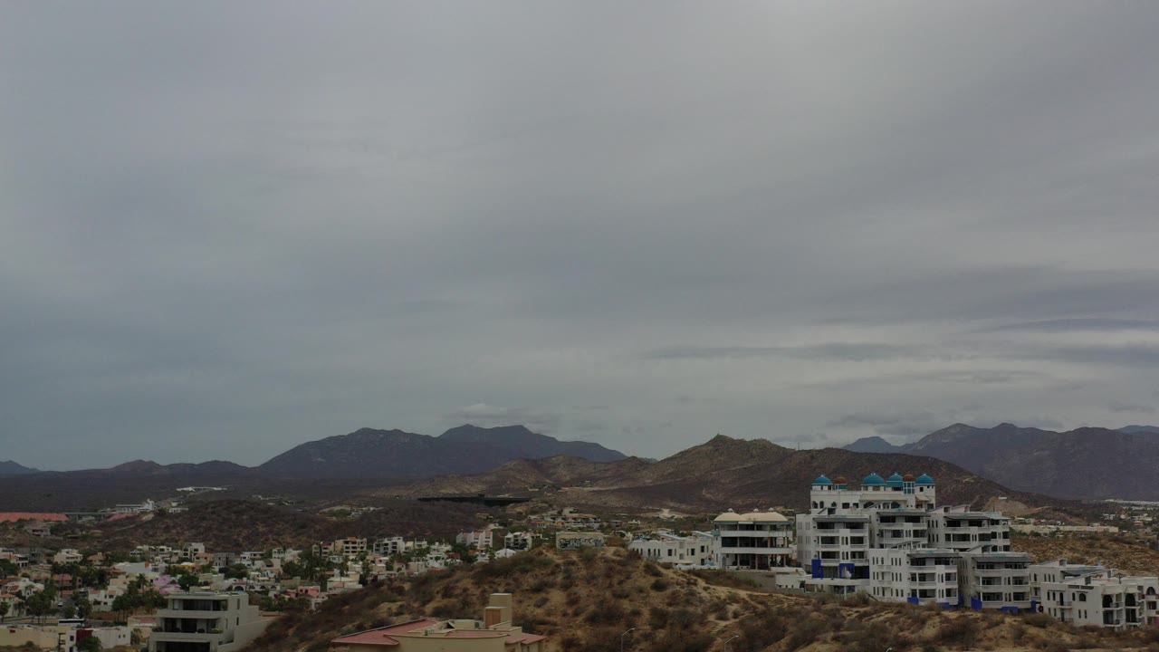 Wide elevated view in Los Cabos, Mexico, showing white hillside buildings, dry scrub slopes, distant dark mountains, and no visible people at the start of the clip.