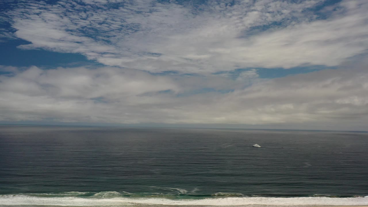Opening frame of the Los Cabos coastline clip with dark green water, a thin white surf band at the bottom, broken clouds above the horizon, a small distant boat, and no visible people.