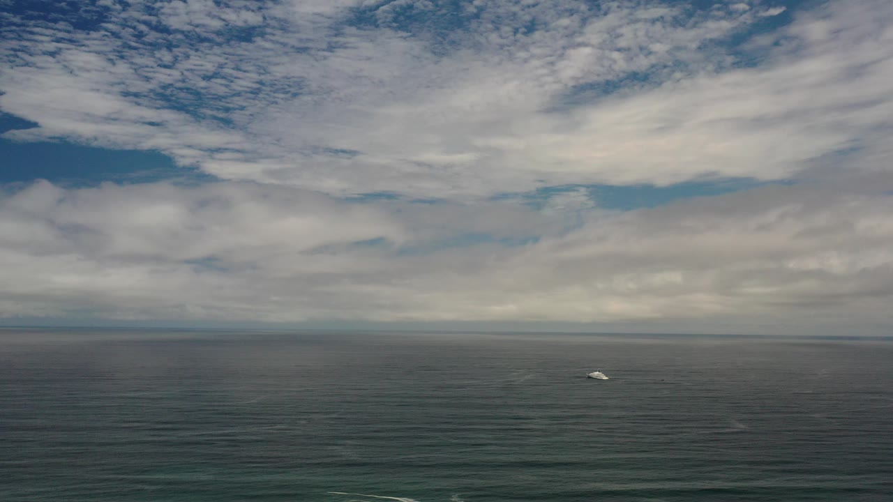 Wide Los Cabos ocean view at the start of the clip, with layered white clouds over blue-gray water, a small distant white boat, and no visible people.