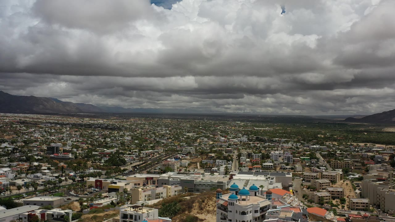 Aerial view of Los Cabos with low-rise urban blocks, a mountain ridge in the distance, gray weather clouds filling the upper frame, and no visible people.