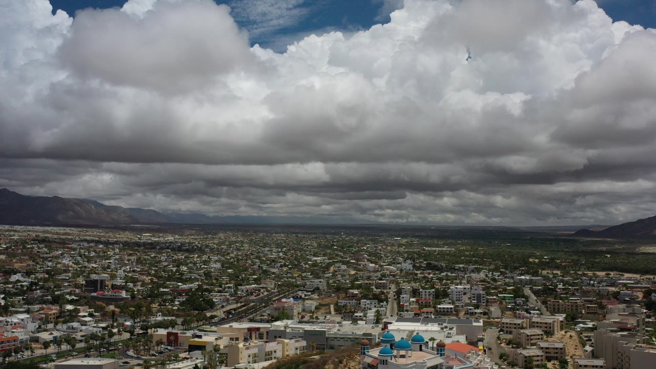 Opening frame of Los Cabos, Mexico from an elevated view, with the city low in frame, distant mountains on the horizon, large clouds overhead, and no visible people.