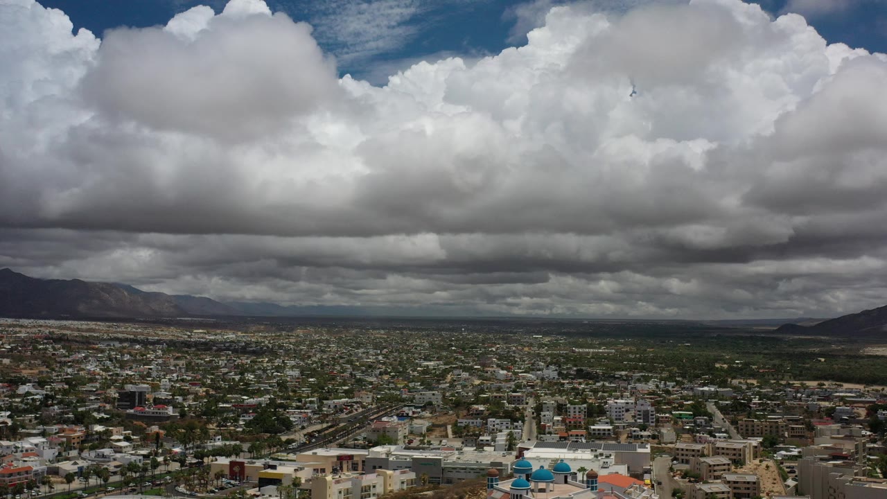 A wide Los Cabos aerial shows cotton-like clouds across the upper frame, a dark cloud band near the horizon, scattered city buildings below, and no visible people.