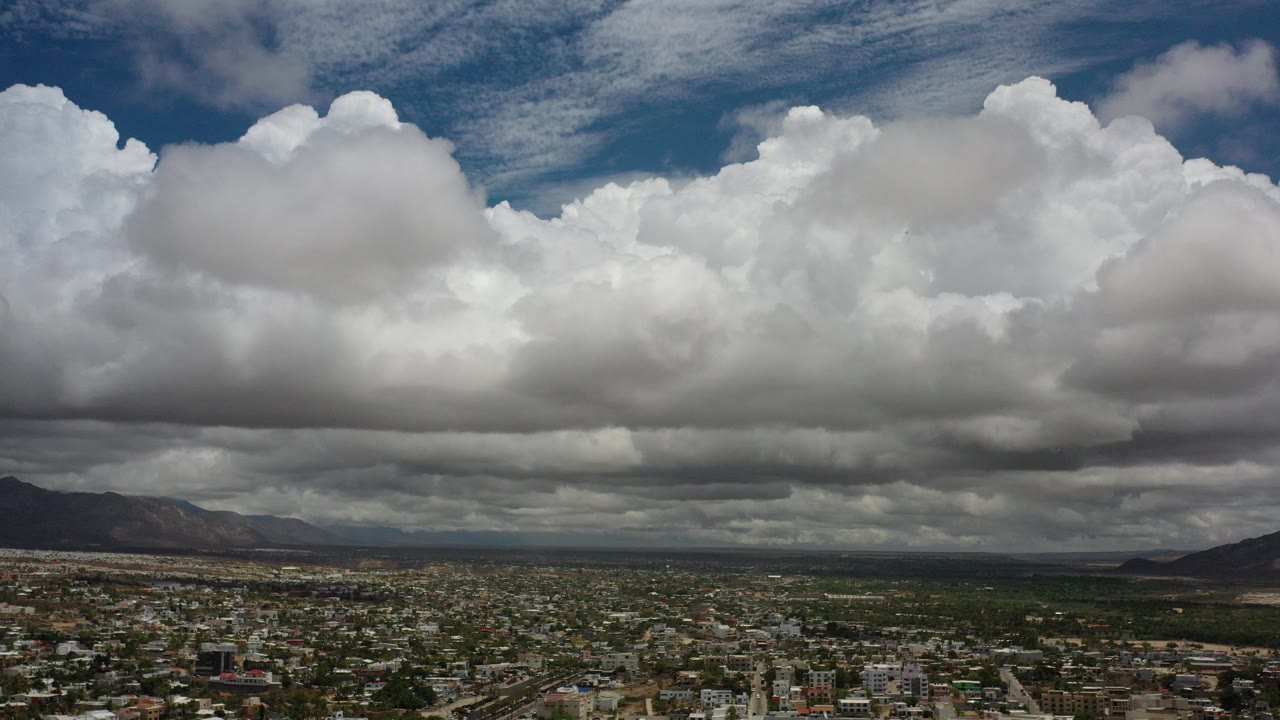 Opening view of Los Cabos, Mexico with layered white and gray clouds above a low cityscape, green terrain, distant mountains, and no visible people.