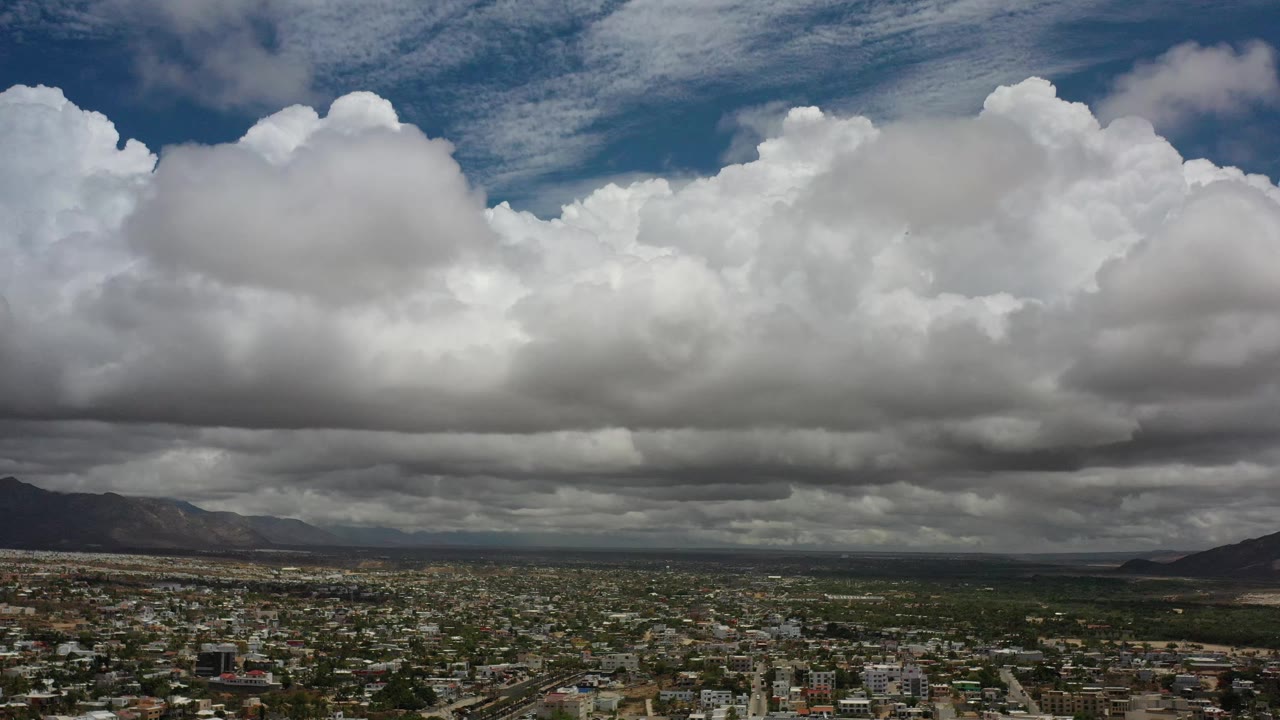 Los Cabos, Mexico appears beneath a broad shelf of gray-bottomed clouds, with a dark mountain mass near the center and no visible people.
