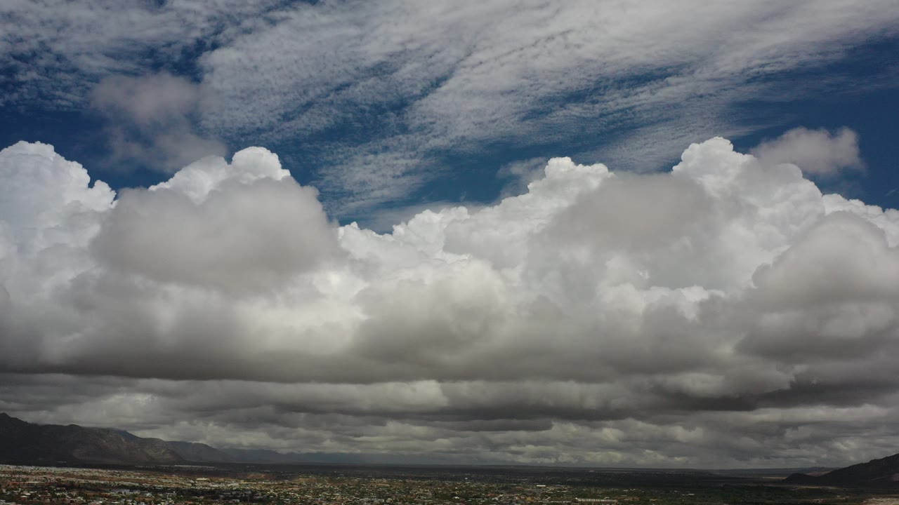 Opening frame of Los Cabos, Mexico with a low desert horizon, distant mountains, towering white clouds, darker cloud bases, blue sky above, and no visible people.