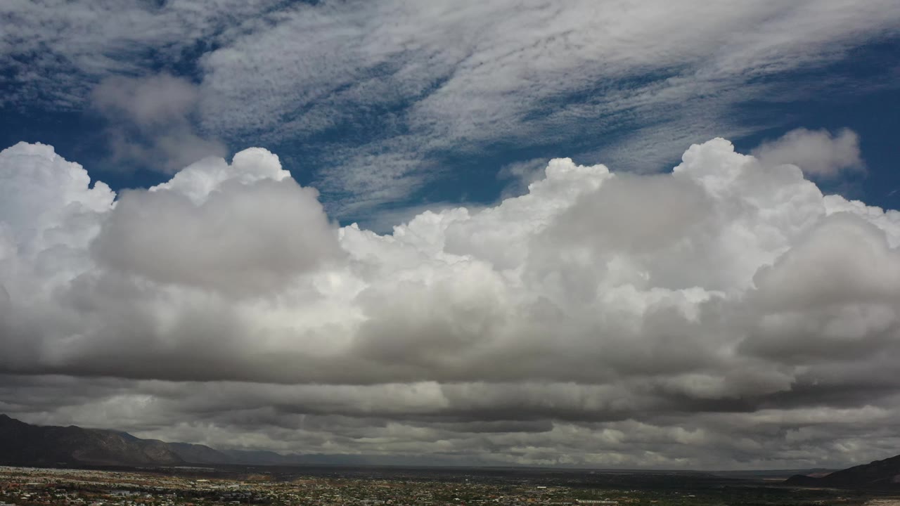 Los Cabos, Mexico sky view with a dense white cloud wall across the center, gray shadowed undersides, a narrow strip of desert terrain, distant mountains, and no visible people.