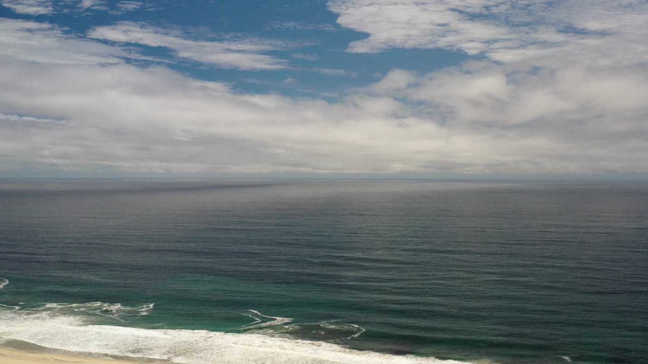 Opening frame of a Los Cabos aerial ocean view with layered clouds, a dark blue-green sea, white surf at the lower edge, and no visible people.