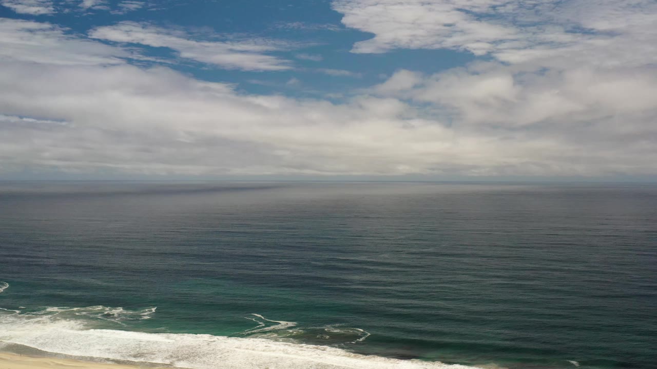 Los Cabos aerial frame with broad cloud bands over the horizon, textured dark water across the center, a bright surf line below, and no visible people.