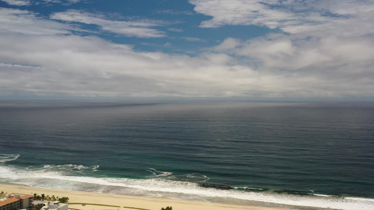 Opening frame of a Los Cabos, Mexico coastline with dark blue water, white surf, pale sand, cloud cover, and no visible people.