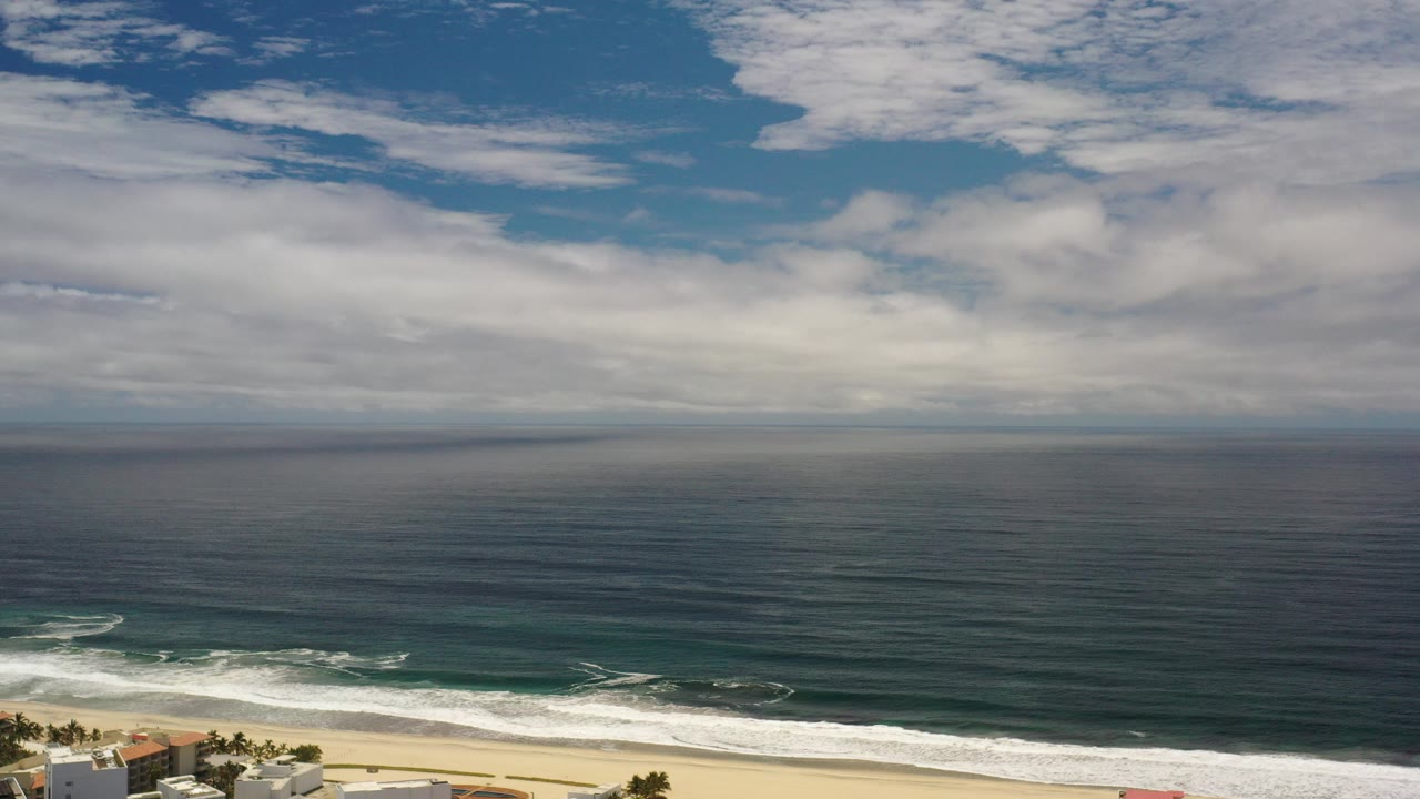 High view over the Los Cabos shoreline with dark ocean water, white surf, pale beach, clouds across the sky, and no visible people.
