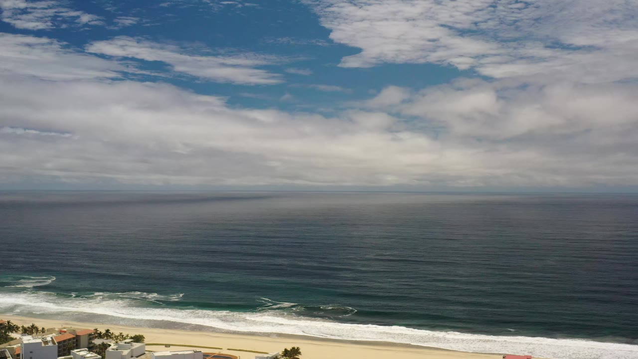Los Cabos ocean view with a broad cloudy sky, dark water bands, breaking waves near shore, and no visible people.